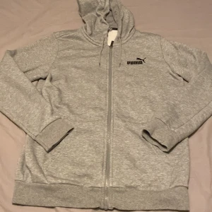 Grå zip hoodie från Puma - Säljer en klassisk grå zip hoodie från Puma med dragkedja framtill och svart logga på bröstet. Tröjan har huva med snörning och två fickor på sidorna. Perfekt för en avslappnad stil eller till träningen. Helt ny bara testad en gång. Pris kan diskuteras och det är bara att höra av er vid frågor😊