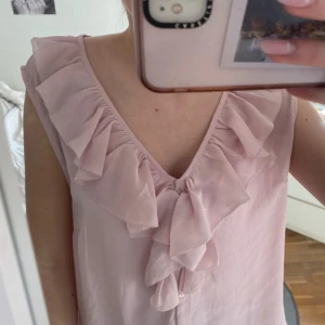 Ljusrosa blus med volang från H&M - Säljer en ljusrosa ärmlös blus från H&M med v-ringning och stora volangdetaljer längs ringningen. Blusen har knappar framtill och är tillverkad i ett lätt, tunt tyg som ger ett luftigt intryck.