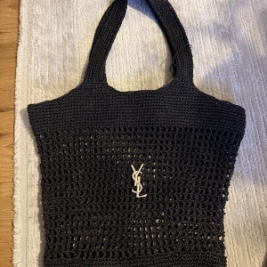 Svart stickad shoppingväska från Yves Saint Laurent - Säljer en svart stickad shoppingväska från Yves Saint Laurent med klassisk YSL-logga i silver framtill. Väskan har ett öppet, nätliknande mönster och breda handtag, perfekt för att bära med sig allt man behöver.