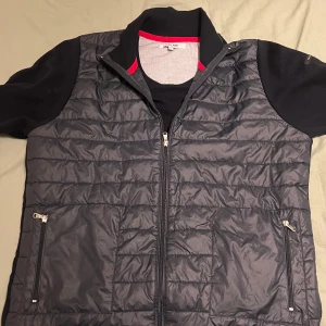 Svart quiltad väst från Iceland - Snygg svart quiltad väst från Iceland med dragkedja framtill och två fickor med dragkedja. Cardiganen har en sportig look och passar perfekt över en tröja för extra värme. Röd detalj på insidan av kragen. XL men passar L