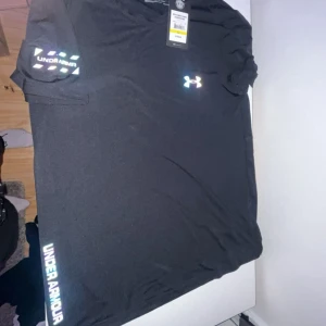 Svart t-shirt från Under Armour - Säljer en svart t-shirt från Under Armour med reflekterande logga på bröstet och ärmen. T-shirten är tillverkad i ett syntetmaterial som andas och har en normal passform. Perfekt för träning eller sportiga aktiviteter.
