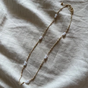 Guldfärgat halsband med pärlor - Ett elegant halsband med tunn guldfärgad kedja och flera vita pärlor jämnt fördelade längs kedjan. Perfekt för att ge en stilren touch till din outfit.