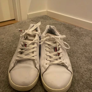 Tommy hilfiger skor! - Säljer ett par vita sneakers från Tommy Hilfiger med klassisk design. Skorna har snörning, rund tå och detaljer i rött och blått på plösen. Yttermaterialet är i skinn och sulan är platt med svart och röd undersida.