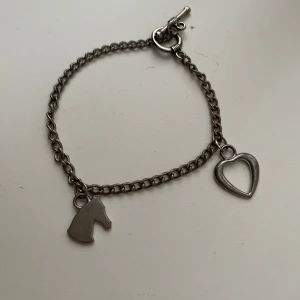 Silverfärgat armband med häst och hjärta - Säljer ett stilrent armband i silverfärg med två berlocker: en häst och ett öppet hjärta. Kedjan är tunn och har ett klassiskt T-lås. Perfekt för dig som gillar hästar eller vill ha en personlig accessoar.