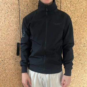 Svart vindjacka från Race Marine - Säljer en svart vindjacka från Race Marine i mycket fint skick, med hög krage och hel dragkedja. Jackan har tryck med Race Marine-logga på ärmen. Perfekt för blåsiga dagar och sportiga outfits.