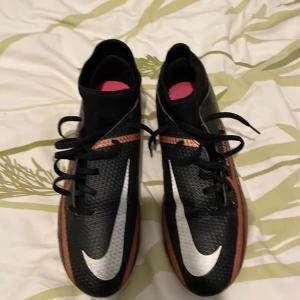 Nike fotbollsskor i svart och koppar - Säljer ett par Nike fotbollsskor i svart med kopparfärgade detaljer och vit sula med rosa dobbar. Skorna har snörning och en stor vit Nike-logga på ovansidan. Perfekta för dig som vill sticka ut på planen!