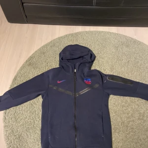 Marinblå Nike FC Barcelona hoodie - Säljer en blå hoodie från Nike med FC Barcelona-logga på bröstet. Hoodiem har dragkedja, huva och två fickor framtill. Perfekt för dig som älskar Barcelona och vill bära deras logga på bröstet.