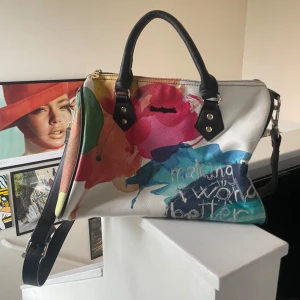 Färgglad handväska från Desigual - Bara använd några gånger, jättebra skick. Nypris ca 800. En unik handväska från Desigual med färgstarkt abstrakt mönster och textdetaljer. Väskan har svarta handtag, avtagbar axelrem och guldfärgade metalldetaljer. Insidan är fodrad i rosa tyg och har en innerficka med dragkedja.