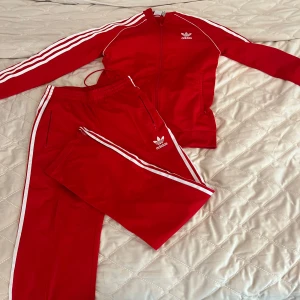 Röda Adidas mjukisset med vita ränder - Säljer ett klassiskt rött Adidas mjukisset med vita ränder längs sidorna och broderad logga framtill. Byxorna har resår i midjan med snörning och är perfekta för en sportig look. Byxorna är i en rak modell. 