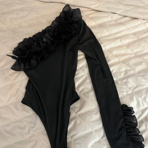 Svart one shoulder body med volang - Säljer en svart body med en ärm och volangdetaljer längs axeln och ärmslutet. Snygg och unik design med oneshoulder och lång ärm. Perfekt för dig som vill sticka ut med en trendig topp.