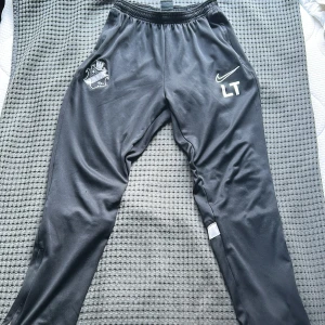 Svarta träningsbyxor från Nike med AIK klubbmärke - Säljer ett par svarta Nike Dri-Fit träningsbyxor med klubbmärke på vänster ben och initialerna LT på höger ben. Byxorna har resår i midjan och är tillverkade i ett syntetmaterial som andas. Perfekta för fotbollsträning eller annan sport.