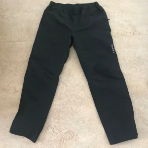 Svarta täckbyxor från Everest - Svarta täckbyxor från Everest med elastisk midja och diskret logga på benet. Perfekta för kalla dagar och utomhusaktiviteter. Byxorna har rak passform och är tillverkade i ett slitstarkt material. Vattentäta. 