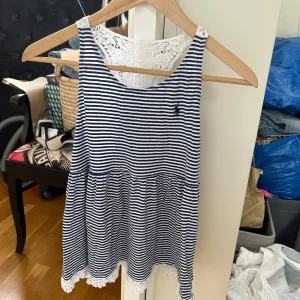 Randigt linne med spets från Polo Ralph Lauren - Säljer en blåvit randig linne från Polo Ralph Lauren i storlek XL (barn) / dvs xs dam. Linnet har ärmlös design, spetsdetaljer upptill och nertill samt rund halsringning. Perfekt för varma dagar med sin luftiga passform och fina spetsdetaljer.