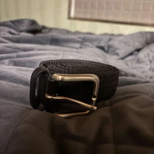 Svart flätat bälte med silverfärgat spänne - Säljer ett svart flätat bälte med ett stilrent silverfärgat metallspänne. Bältet har en klassisk design och passar perfekt till jeans eller chinos. Detaljer i svart läder vid spännet ger en snygg kontrast.