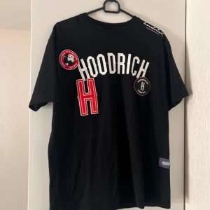 Svart t-shirt från Hoodrich med patchar - Svart t-shirt från Hoodrich med stora vita bokstäver och röda detaljer framtill. Flera broderade patchar och tryck, bland annat en stor röd H och rund logga på bröstet. Klassisk passform och rund halsringning.