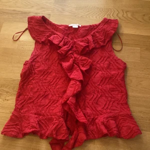 Röd volangblus från Gina Tricot - Säljer en röd blus från Gina Tricot med volangdetaljer framtill och längs nederkanten. Blusen har ett strukturerat tyg och är ärmlös, perfekt för att ge outfiten lite extra edge.