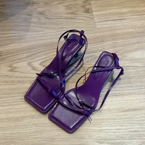 Lila klackar med smala remmar och klack - Säljer ett par trendiga lila sandaler från Zara med smala remmar och fyrkantig tå. Skorna har en lagom klack och tunna band som går runt foten. Aldrig använda. Litet märke under foten från butiken som inte syns alls.
