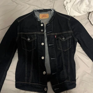 Mörkblå jeansjacka från Levi's - Klassisk mörkblå jeansjacka från Levi's med silverfärgade knappar och två bröstfickor med lock. Snygga kontrastsömmar och rak passform. Perfekt att slänga över en hoodie eller t-shirt.