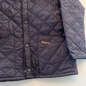Svart quiltad jacka från Barbour - Klassisk svart quiltad jacka från Barbour med knäppning framtill och två stora fickor. Jackan har en bred krage i manchester och Barbour-logga broderad på fickan. Den är i storleken M (barnstorlek) vilket motsvarar ungefär XSS (herrstorlek). Nypris: ca 1700 kr och mitt pris: 530 kr. Perfekt för kyliga dagar och tidlös stil. Skriv bara om du har frågor, priset kan diskuteras!
