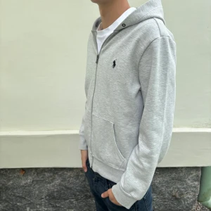 Polo Ralph Lauren zip - Säljer en stilren ljusgrå hoodie från Polo Ralph Lauren med dragkedja och huva. Tröjan har två fickor framtill och en liten broderad logga på bröstet. Perfekt för en avslappnad look. Tyvärr inga snören. 