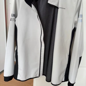 Vit och svart vindjacka från Sail Racing - Snygg vit och svart vindjacka från Sail Racing med dragkedja framtill och på fickor. Jackan har lång ärm, sportig design och tryckt logga på ärmen. Perfekt för dig som gillar stilren och funktionell ytterjacka. Jackan är liten i storleken. 