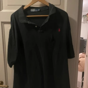 Svart pikétröja från Polo Ralph Lauren - Klassisk svart pikétröja från Polo Ralph Lauren med korta ärmar och röd broderad logga på bröstet. Skön passform och mjukt bomullsmaterial, perfekt till många olika outfits.
