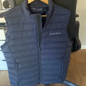 Mörkblå dunväst från J.Lindeberg - Snygg mörkblå dunväst från J.Lindeberg med diskret logga på bröstet. Västen har dragkedja framtill och två fickor. Perfekt för lager-på-lager och ger en sportig look.