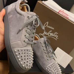 Gråa sneakers med nitar från Christian Louboutin - Säljer ett par gråa sneakers från Christian Louboutin i mocka med silverfärgade nitar på tån. Skorna har klassisk snörning och den ikoniska röda sulan. Perfekta för dig som vill sticka ut med en lyxig och edgy stil.