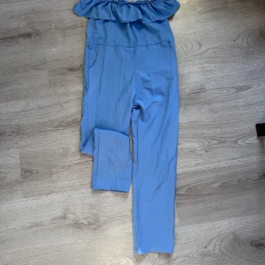 Ljusblå byxdress med volang och bälte stl 140 - Säljer en ljusblå byxdress med bred volang upptill och tillhörande bälte i midjan. Byxdressen har raka ben och är tillverkad i ett mjukt, lätt material. Perfekt för dig som vill ha en stilren och trendig look.