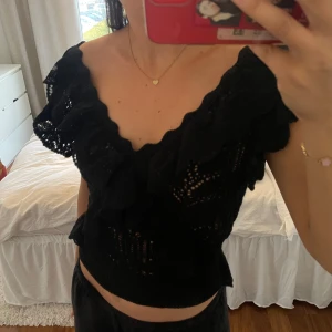 Svart volangtopp med djup v-ringning - Säljer en svart topp med djup v-ringning och volangdetaljer längs axlar och halsringning. Toppen är ärmlös och har en croppad passform, perfekt för att lyfta en outfit med lite extra edge.
