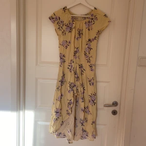 Gul blommig klänning från Lindex - Söt gul klänning från Lindex med lila och lila-blå blommor. Klänningen har korta ärmar, rund halsringning och shorts med ett fint släp bakom . Perfekt för sommarens varma dagar och har en fin, solig känsla.