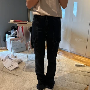 Mörka cargo jeans från Zara - Säljer ett par mörka jeans från Zara med raka ben och flera fickor på sidorna. Byxorna har normal passform och stängs med knapp och dragkedja. Perfekta för en avslappnad och trendig stil. Låg midja och inte använda många gånger pga för små, storlek 32! (168 cm lång för referens)
