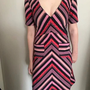 Rosa och svart randig klänning med v-ringning - Säljer en snygg klänning med djup v-ringning och korta ärmar. Klänningen har ett randigt mönster i rosa, svart och rött med markerad midja och fickor framtill. Perfekt för dig som gillar färg och mönster!