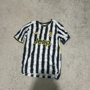 Juventus fotbollströja Pogba #10 - Juventus matchtröja med svart och vitt randigt mönster, gula detaljer och Pogba #10 på ryggen. Jeep-logga på framsidan och Serie A-märke på ärmen. Klassisk V-ringning och kortärmad modell.