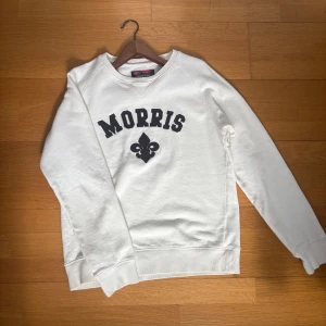 Vit tröja från Morris - Säljer en vit tröja från Morris med svart tryck och dekorativt emblem framtill. Tröjan har rund halsringning och långa ärmar. Perfekt för en avslappnad och stilren look.