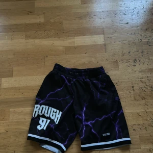 Svarta basket shorts med lila blixtmönster från Black Squad - Coola svarta shorts från Black Squad med lila blixtar och vit text 'ROUGH 91' på ena benet. Elastisk midja och vita detaljer längs bensluten. Perfekta för en sportig och avslappnad stil.