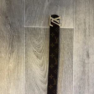 Brunt bälte med monogram och guldfärgat spänne - Säljer ett brunt bälte med klassiskt monogrammönster och ett stort Bältet har en stilren design och är tillverkat i skinn med tydliga logodetaljer längs hela remmen.