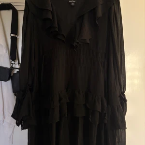 Svart volangklänning från Boohoo - Svart långärmad klänning från Boohoo med volangdetaljer längs ärmar, nederkant och krage. Klänningen har snören som gör så att man kan dra åt på sidorna så de blir en mer figursydd passform. Aldrig använd, dock ingen prislapp kvar 