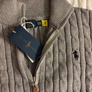Grå kabelstickad tröja från Ralph Lauren - Säljer en snygg grå kabelstickad tröja från Ralph Lauren med halv dragkedja och broderad logga på bröstet. Tröjan har hög krage och ribbade muddar. Perfekt för lager-på-lager och stilren look.