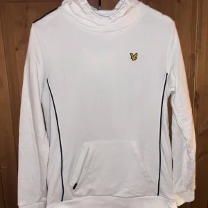 Vit hoodie från Lyle & Scott med svarta detaljer - Vit hoodie från Lyle & Scott med svart piping längs sidorna och axlarna. Klassisk gul logga på bröstet och stor ficka framtill. Mjuk bomullskänsla och huva för extra komfort.  Använd en del men håller fortfarande ett bra skick!