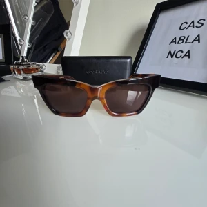  solglasögon Grace från Ace & Tate - Snygga solglasögon från Ace & Tate i modellen Grace, färg caramel blitz. De har en rektangulär form med bred båge och mörka glas. Bågarna är i brunt transparent acetat med en cool, modern look.