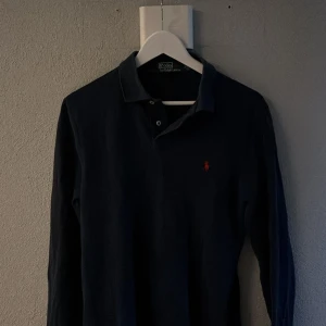 Mörkblå pikétröja från Polo Ralph Lauren - Säljer en klassisk mörkblå långärmad pikétröja från Polo Ralph Lauren. Tröjan har slim fit-passform och den sköna röda loggan broderad på bröstet. Perfekt för en stilren och tidlös look. 10/10 skick!
