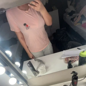 Ljusrosa t-shirt med tryck - Ljusrosa t-shirt med ett litet tryck av en palm och sol på vänster bröst. Rund halsringning och korta ärmar. Perfekt för en avslappnad stil.