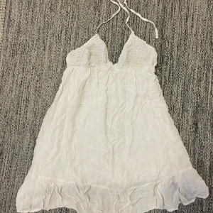 Vit kortklänning med spets och volang - Söt vit kortklänning från Pull&Bear med tunna axelband som knyts i nacken. Klänningen har spetsdetaljer upptill och volang nertill, samt en lätt och luftig känsla. Den är utsvängd och har dubbla tyger. På bild tre kan man se en liten fläck men den syns inte när man väl har på sig klänningen☺️