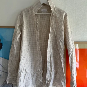 Beige och vit rutig skjorta från Massimo Dutti - Snygg beige och vit rutig skjorta från Massimo Dutti. Skjortan har långa ärmar och klassisk krage, perfekt för en stilren look. Tillverkad i mjuk bomull och stängs med knappar framtill.