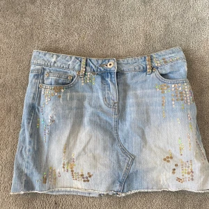 Ljusblå jeanskjol med paljetter - Säljer en ljusblå jeanskjol med rå kant och dekorativa paljetter i silver och holografiska färger framtill och baktill. Kjolen har klassisk femficksdesign och knappstängning. Perfekt för dig som vill sticka ut lite extra!