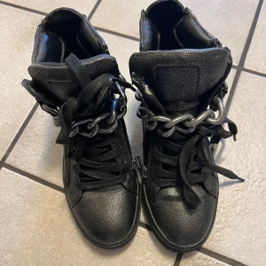 Svarta sneakers med kedjedetalj - Säljer ett par svarta sneakers med grov kedja framtill och dragkedja på sidan. Skorna har snörning, spänne och är tillverkade i syntetmaterial med en platt sula. Perfekta för dig som gillar en edgy stil.