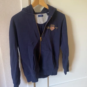 Mörkblå hoodie från Gant - Säljer en mörkblå hoodie från Gant med dragkedja och huva. Tröjan har Gant-logga broderad på bröstet och två fickor framtill. Perfekt för en avslappnad stil.