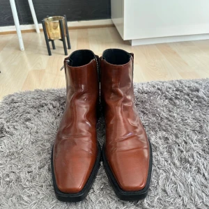 Bruna boots med dragkedja och fyrkantig tå - Snygga bruna boots i skinn med dragkedja på sidan och fyrkantig tå. Skorna har en platt sula och stilren design som passar till många outfits. Perfekta för dig som gillar klassiska men trendiga skor.