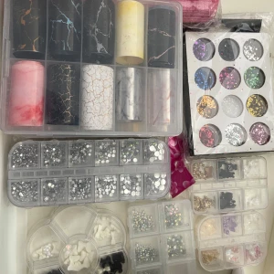 Nageldekorationer och glitter i burkar och askar - Ett set med olika nageldekorationer, inklusive glitter i flera färger, folie i metallic och marmor-mönster samt små stenar och pärlor. Perfekt för att skapa kreativa nageldesigner. Kommer i praktiska förvaringsaskar och burkar.